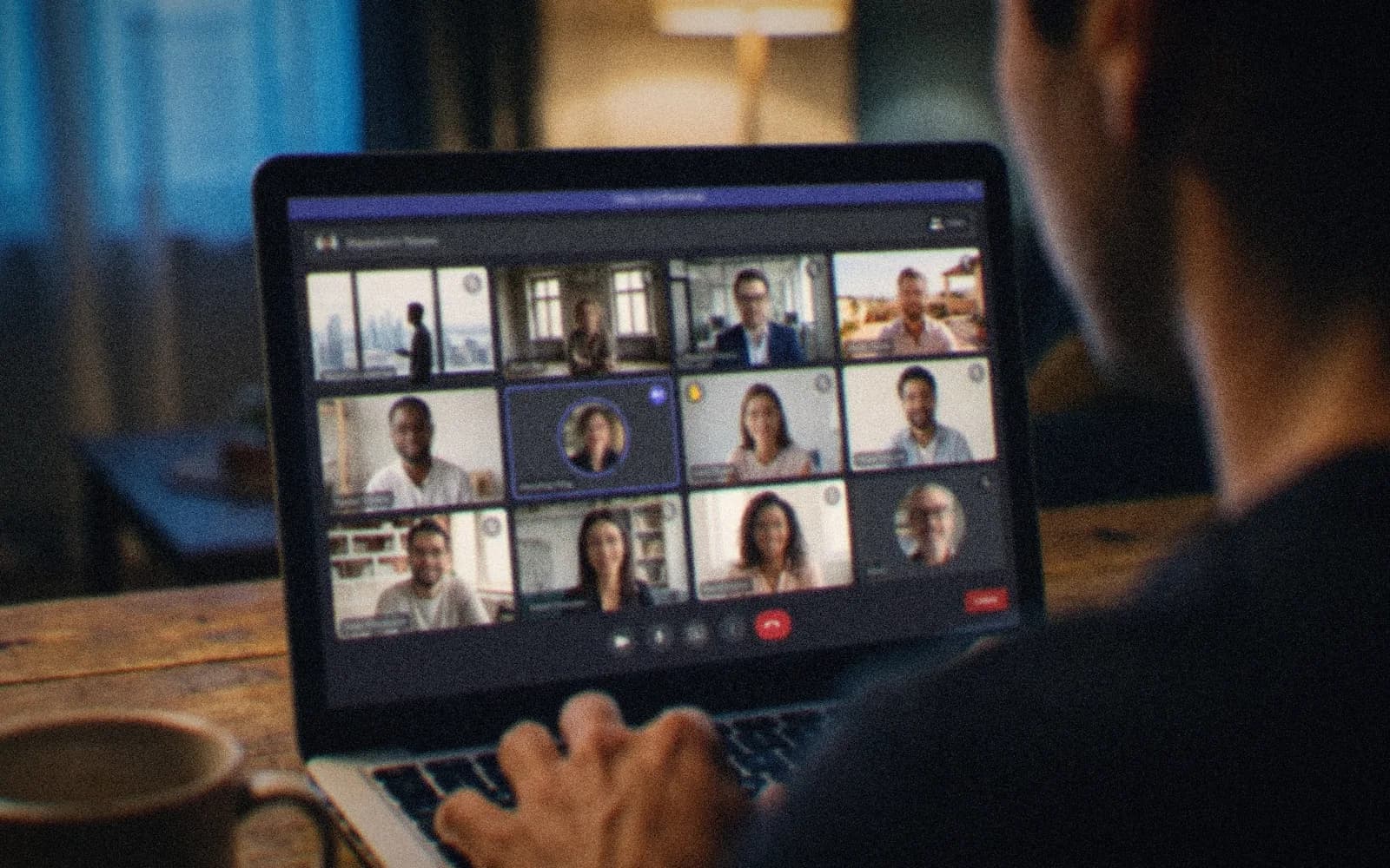 Over the shoulder of someone on a company-wide video call, a gallery of colleagues from several offices on the laptop screen