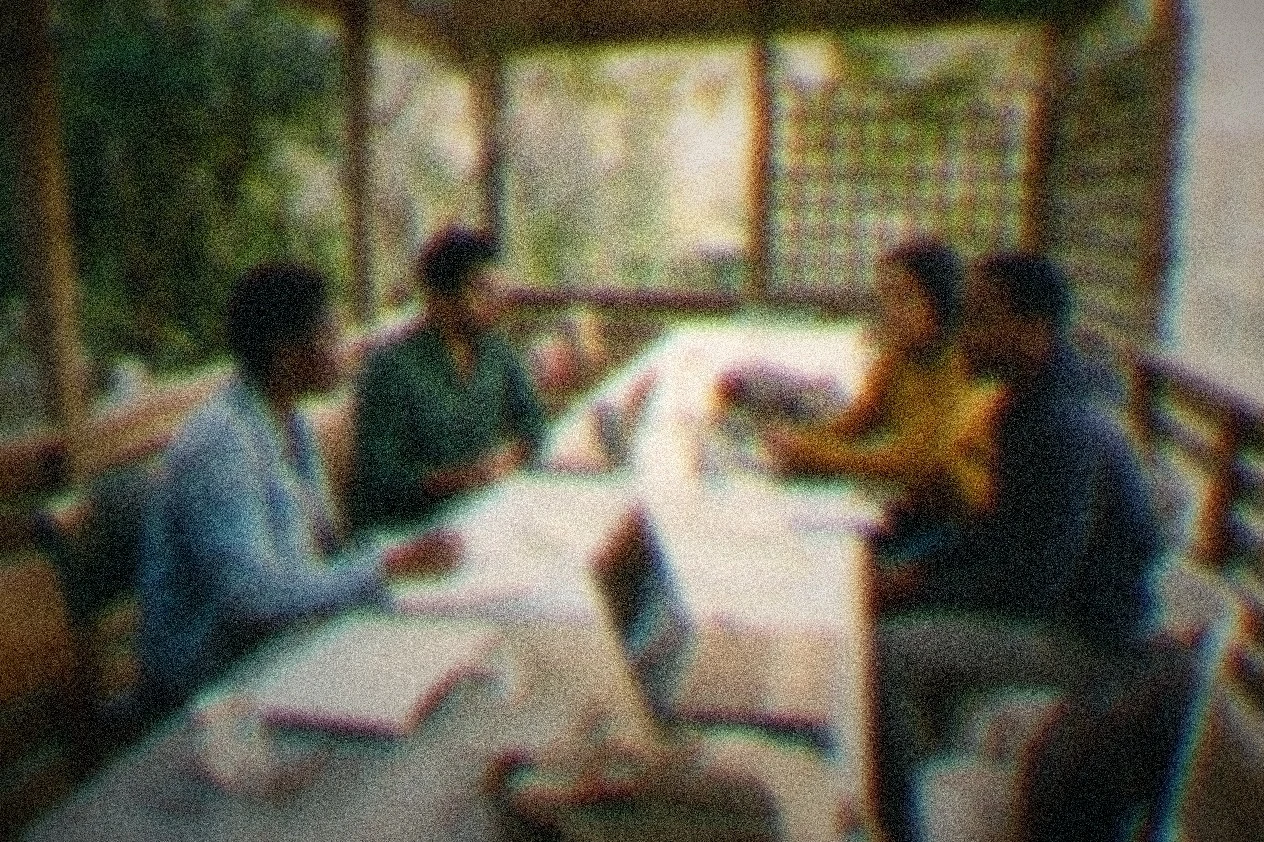 Four people in two small clusters facing each other across a long table of maps and laptops at golden hour.